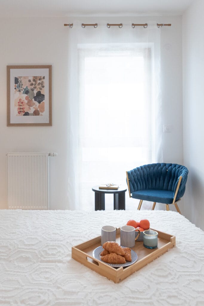 Chambre parentale d’un appartement à Nancy avec fauteuil bleu, mobilier en bois et plateau petit-déjeuner